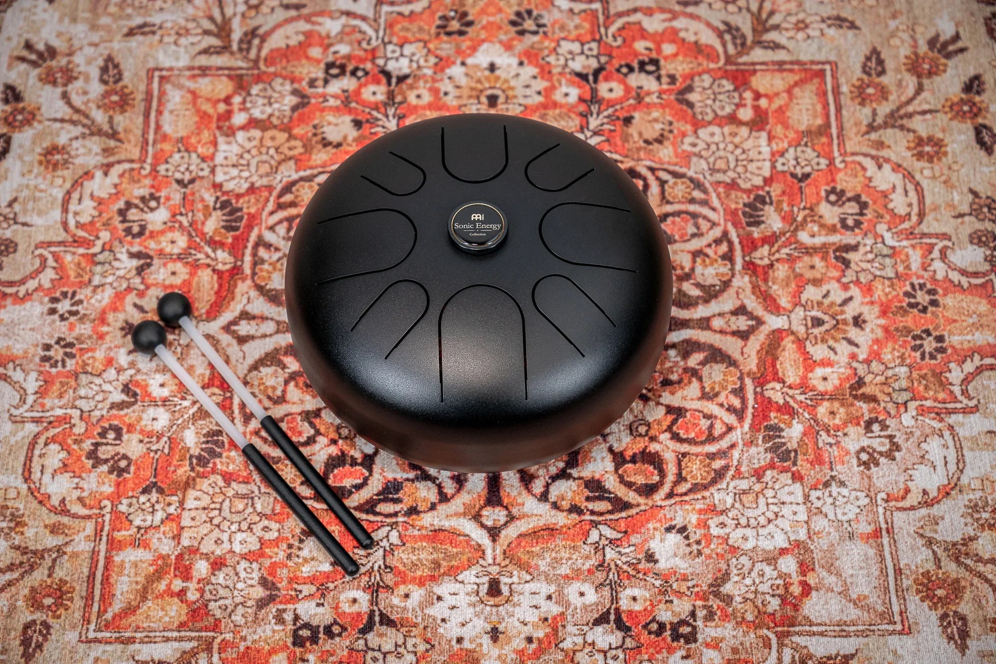 Black steel tongue drum with mallets on a patterned rug, sound healing instrument