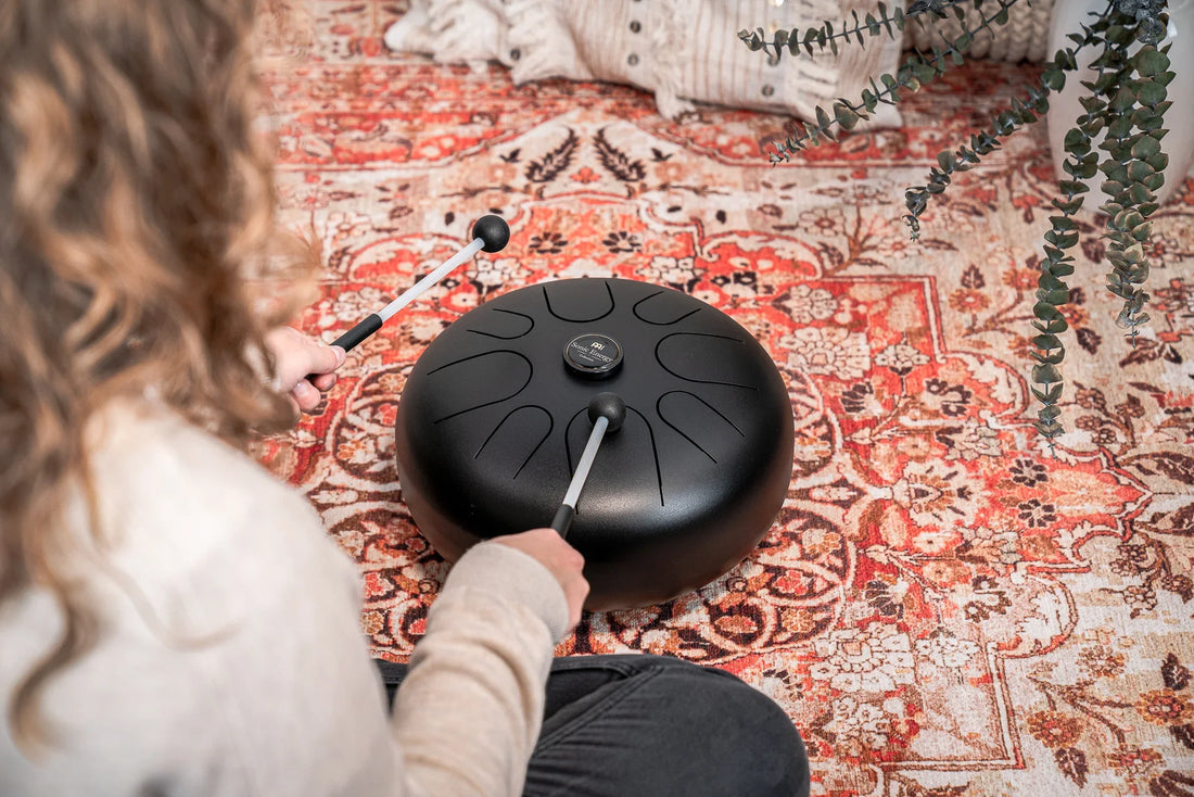Person playing a black steel tongue drum with mallets on a patterned rug