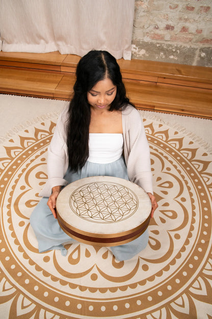 Woman meditating with a hand drum featuring a sacred geometry design, seated on a mandala rug