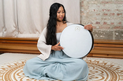 Woman holding a white frame drum for sound healing, seated on a mandala rug indoors
